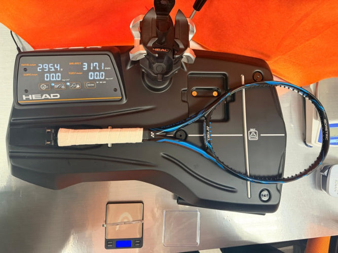A Yonex tennis racquet with a light wood handle and blue/black frame is displayed on a black HEAD racket measurement and fitting machine with a digital display screen, set against an orange background.